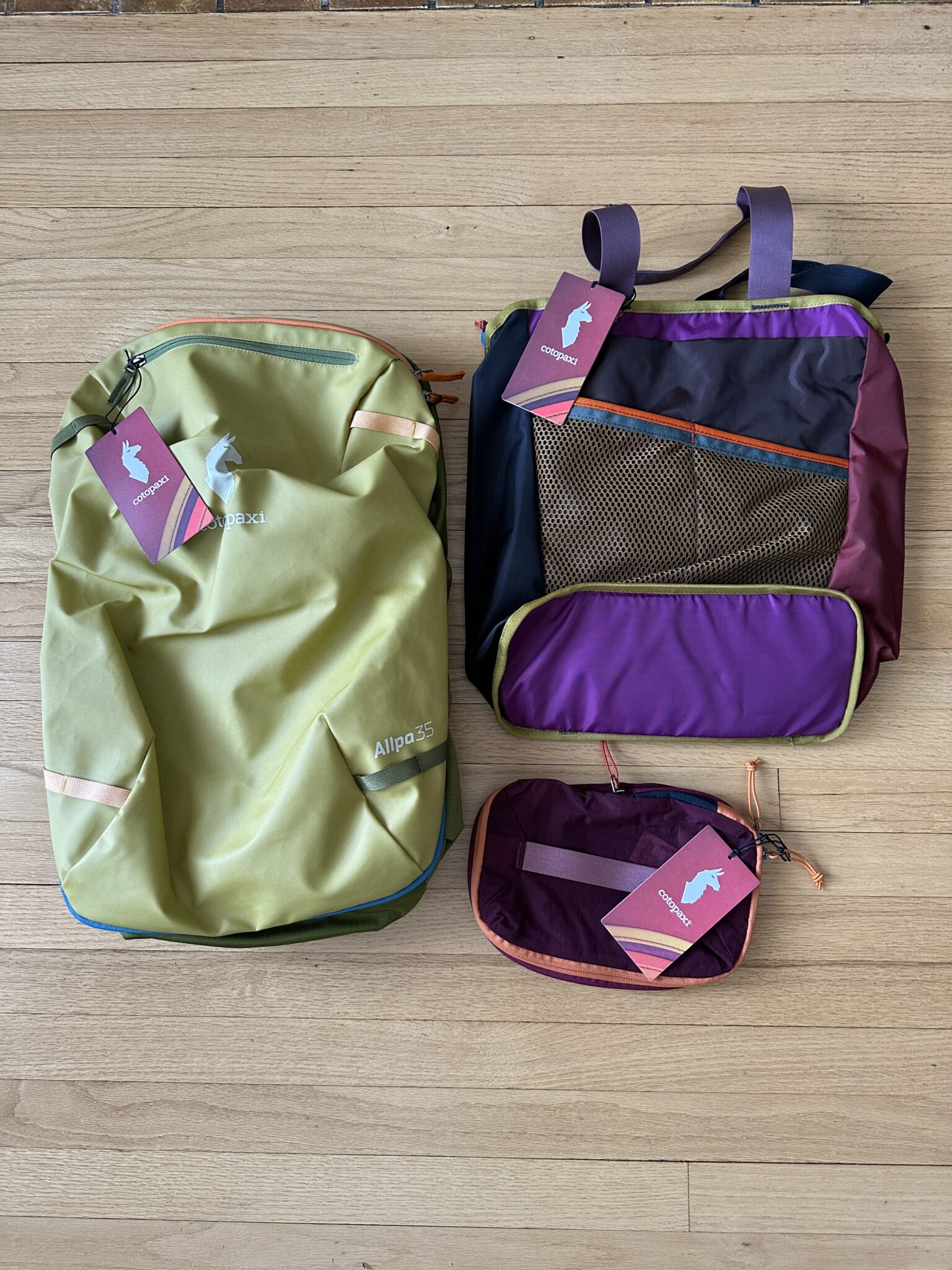 Three Cotopaxi travel bags in green, purple, and multicolor are displayed on a wooden floor, each with a pink tag attached.