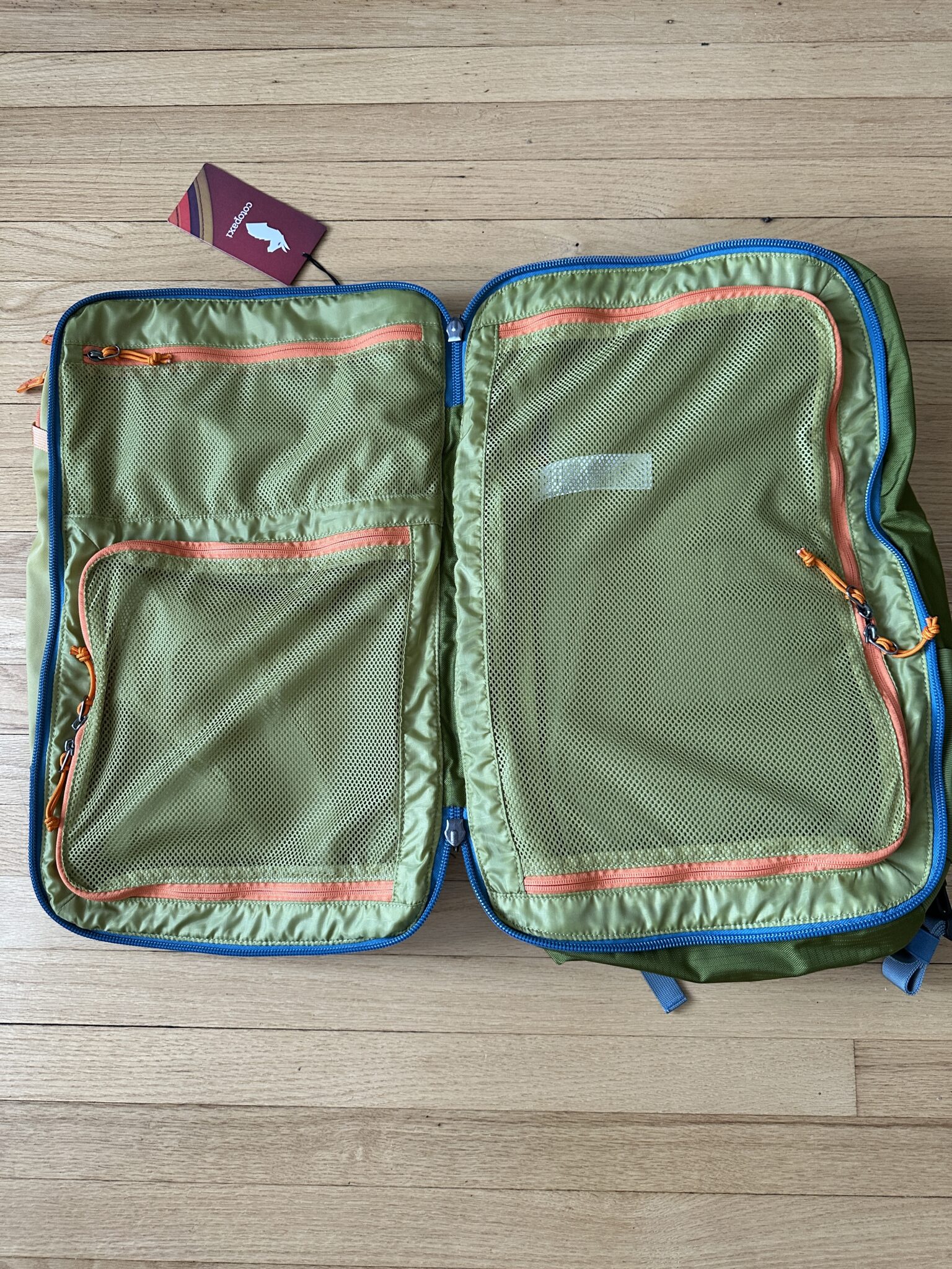 An empty green suitcase with orange zippers is open on a wooden floor; a closed red passport is placed above the suitcase.