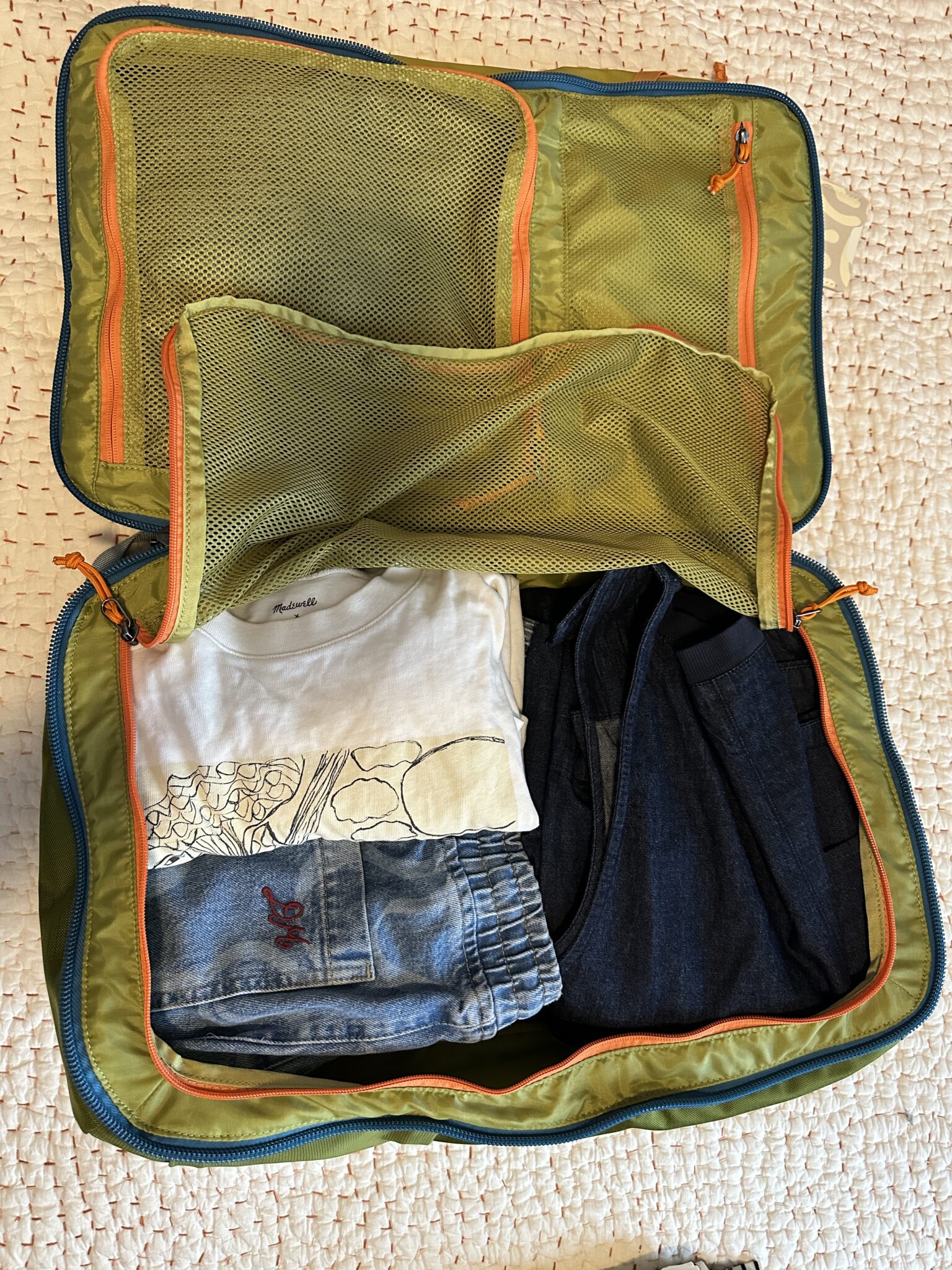 Open green packing cube containing neatly folded clothes, including a white graphic t-shirt, blue jeans, and blue shorts, placed on a textured surface.