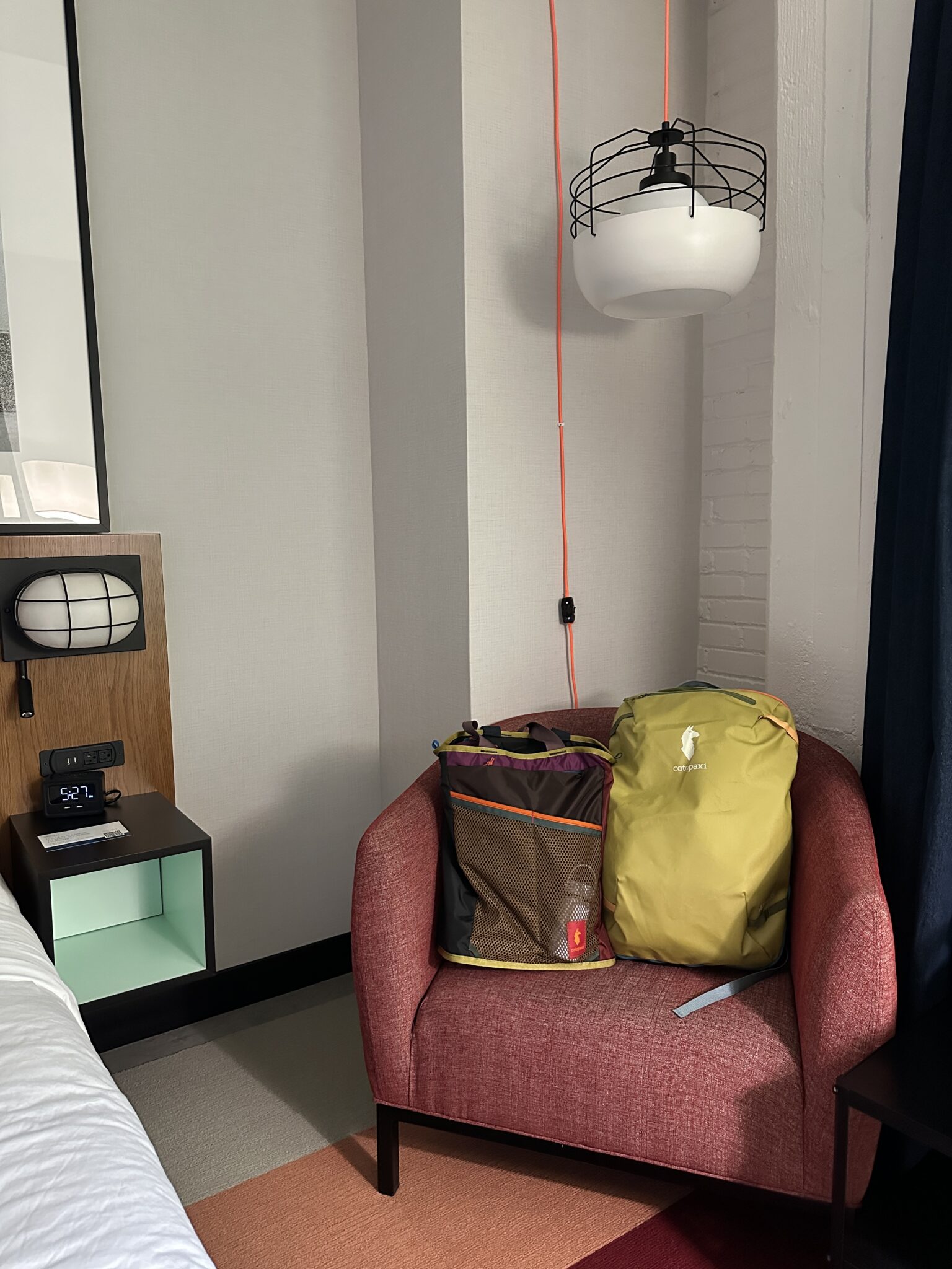 A red armchair with two bags sits next to a nightstand and bed in a hotel room. A wall lamp hangs above, and a digital clock reads 9:21.