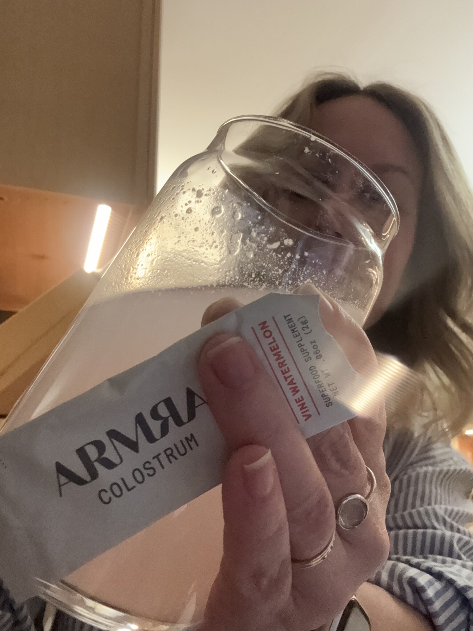 A person holding a glass of liquid and a packet labeled "ARMRA Colostrum WAVE WATERMELON." The packet appears partially submerged in the glass.