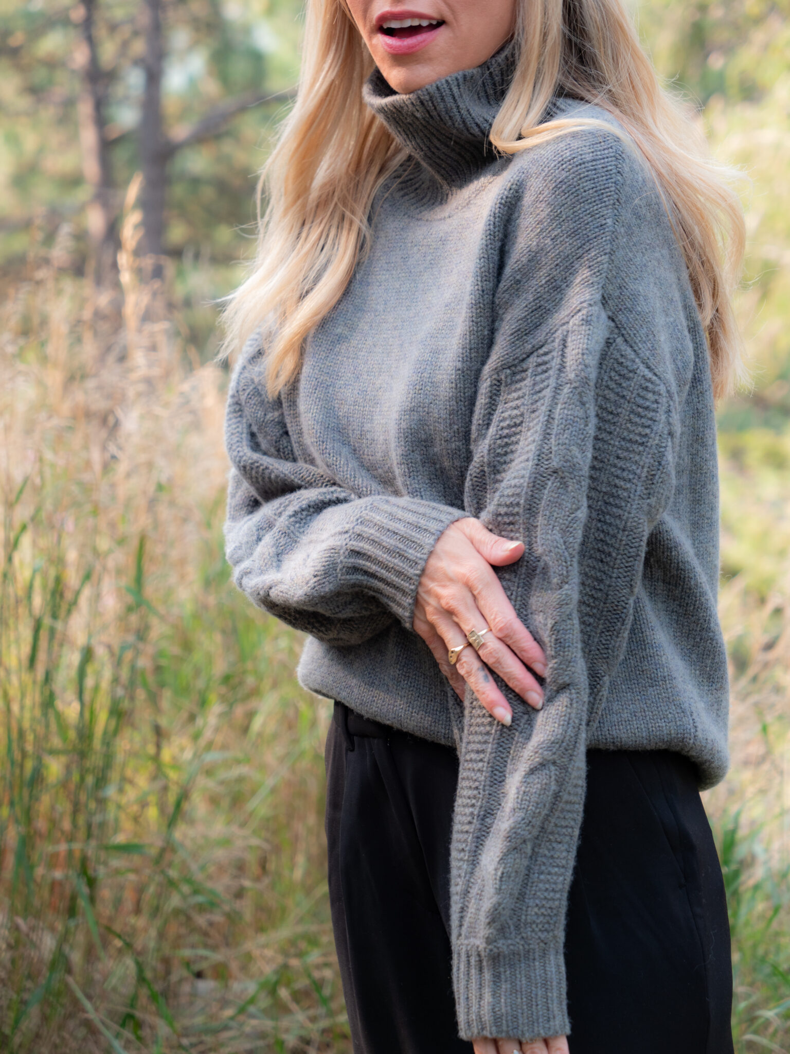 A woman with long blonde hair wearing a gray cable-knit turtleneck sweater and black pants stands outdoors in a grassy area.