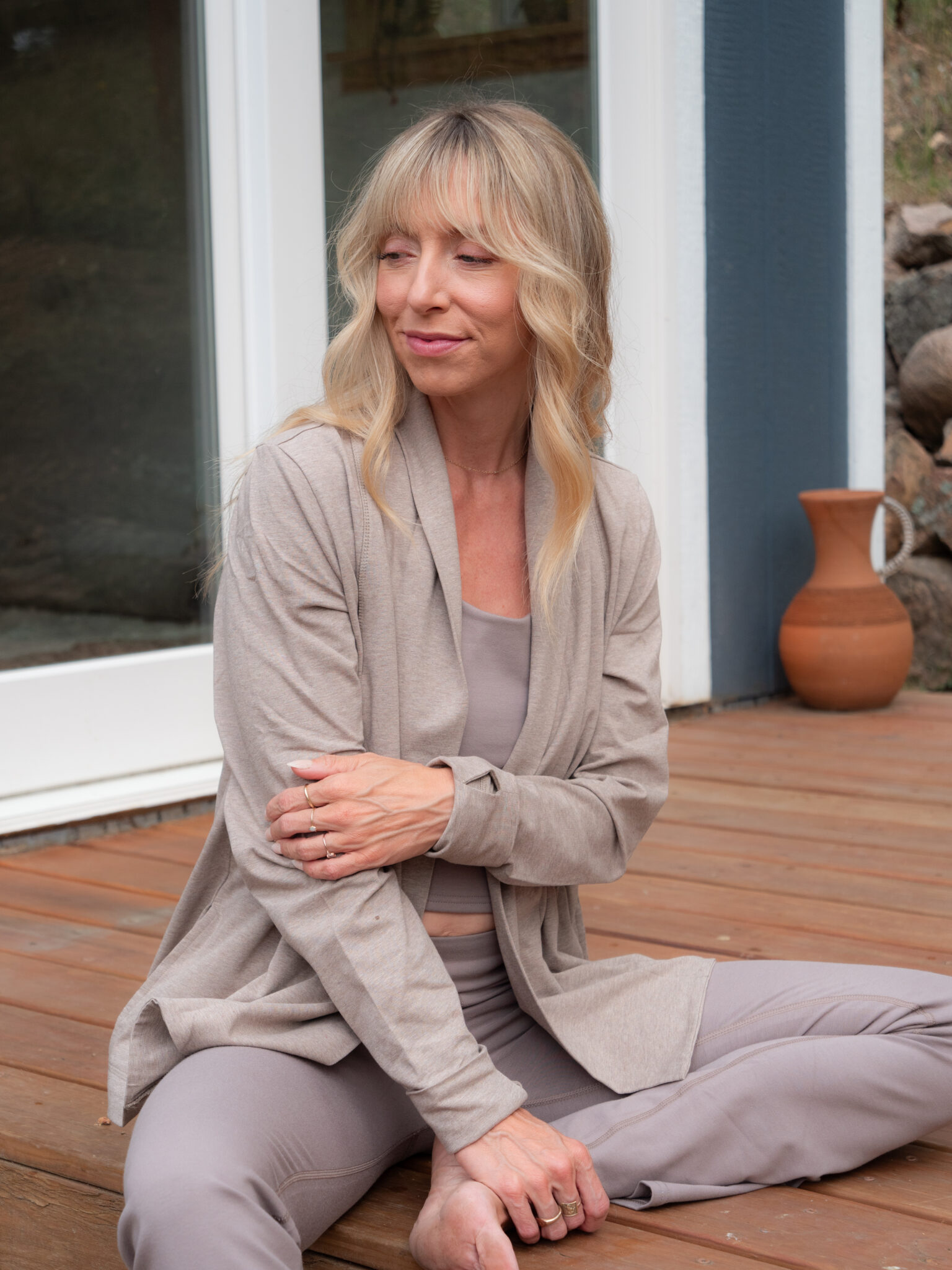 A woman with blonde hair sits cross-legged on a wooden deck, wearing a light beige outfit, with a clay vase in the background.