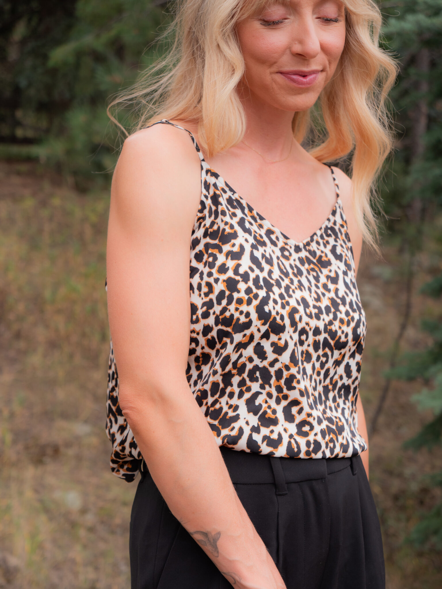 A woman with long blonde hair wears a leopard print sleeveless top and black pants, standing outdoors with trees in the background.