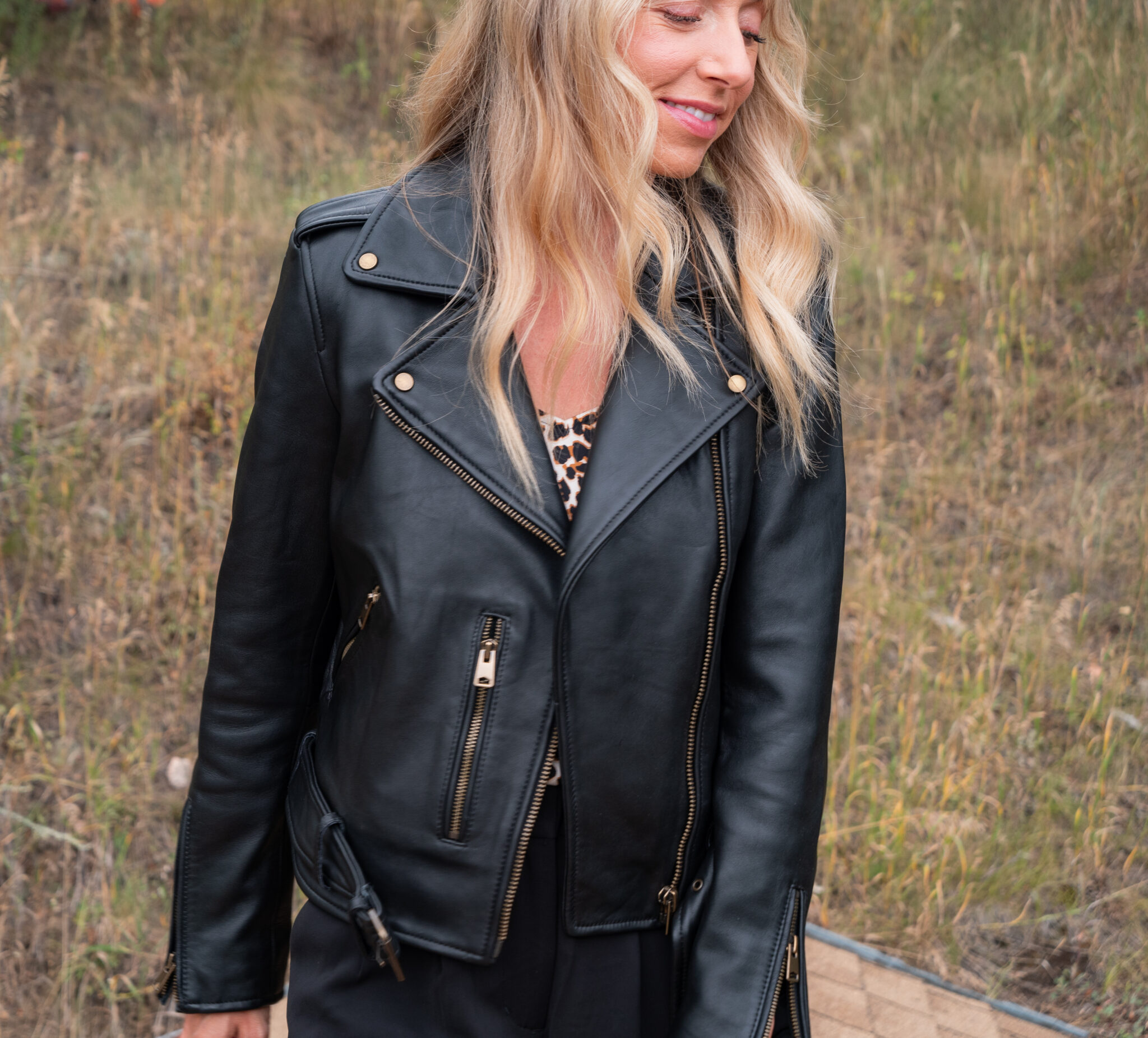 Woman standing outdoors wearing a black leather jacket over a leopard print top, with wavy blonde hair, against a background of dry grass.