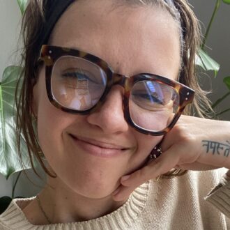 Person wearing large tortoiseshell glasses and a beige sweater smiles with their chin resting on their hand; a "Namaste" tattoo is visible on their wrist.