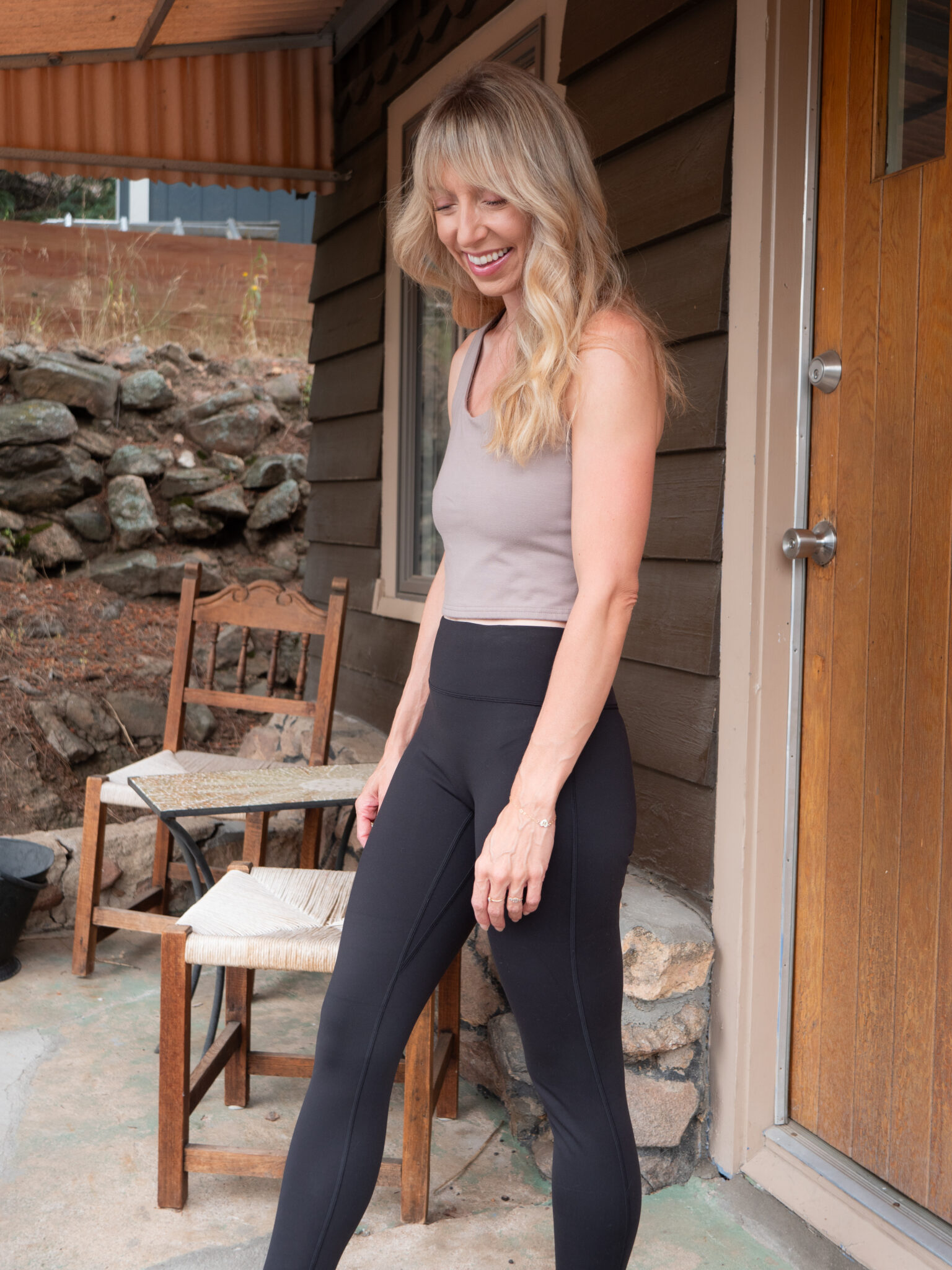 A woman with long blond hair wearing a taupe tank top and black leggings stands outside near a wooden door and two chairs.