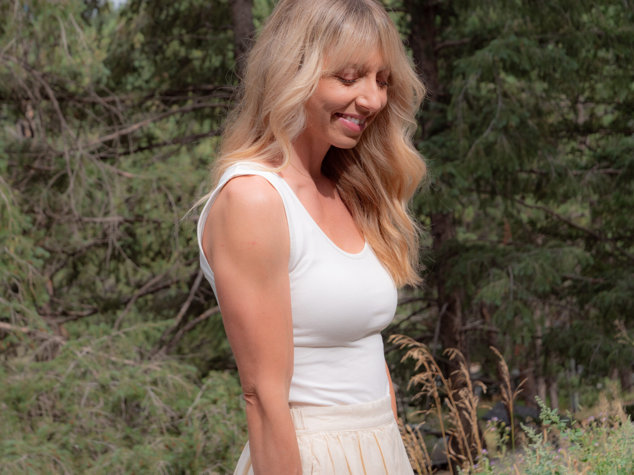 A woman with long blonde hair wearing a white sleeveless top and cream skirt stands outdoors among trees, smiling with eyes closed.
