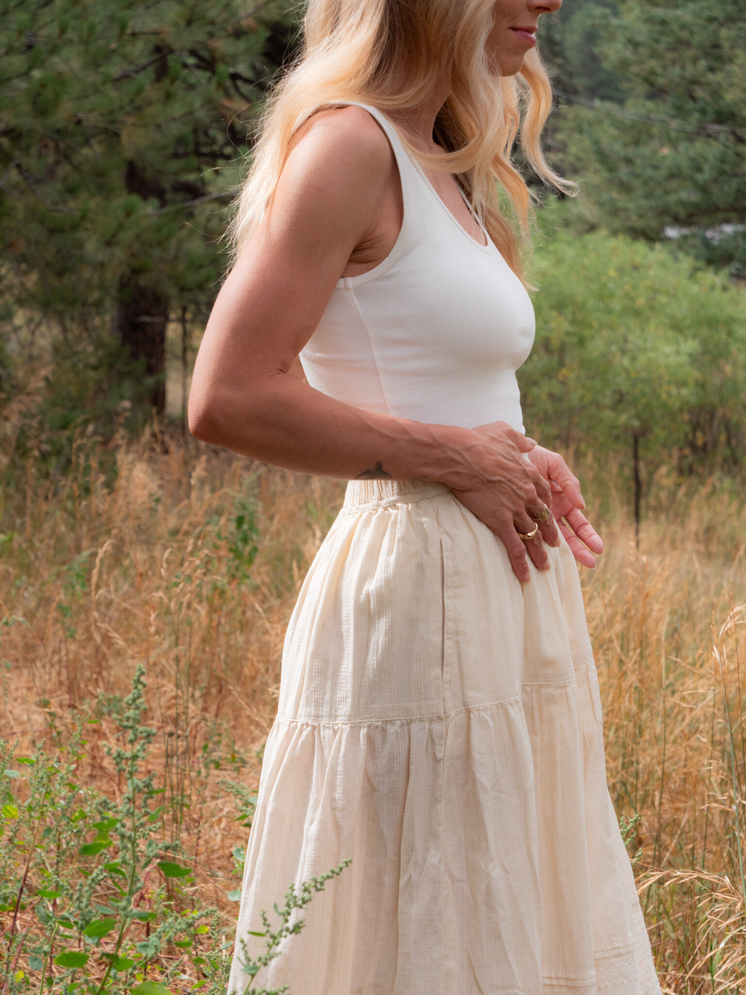 A woman wearing a white tank top and cream skirt stands outdoors in a grassy area with trees in the background.