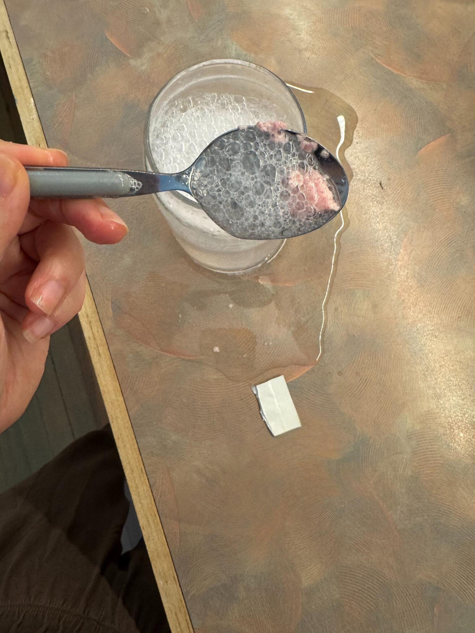 A hand holds a spoon with soapy water and foam above a glass filled with bubbles; water and a small white object are on a brown tabletop.