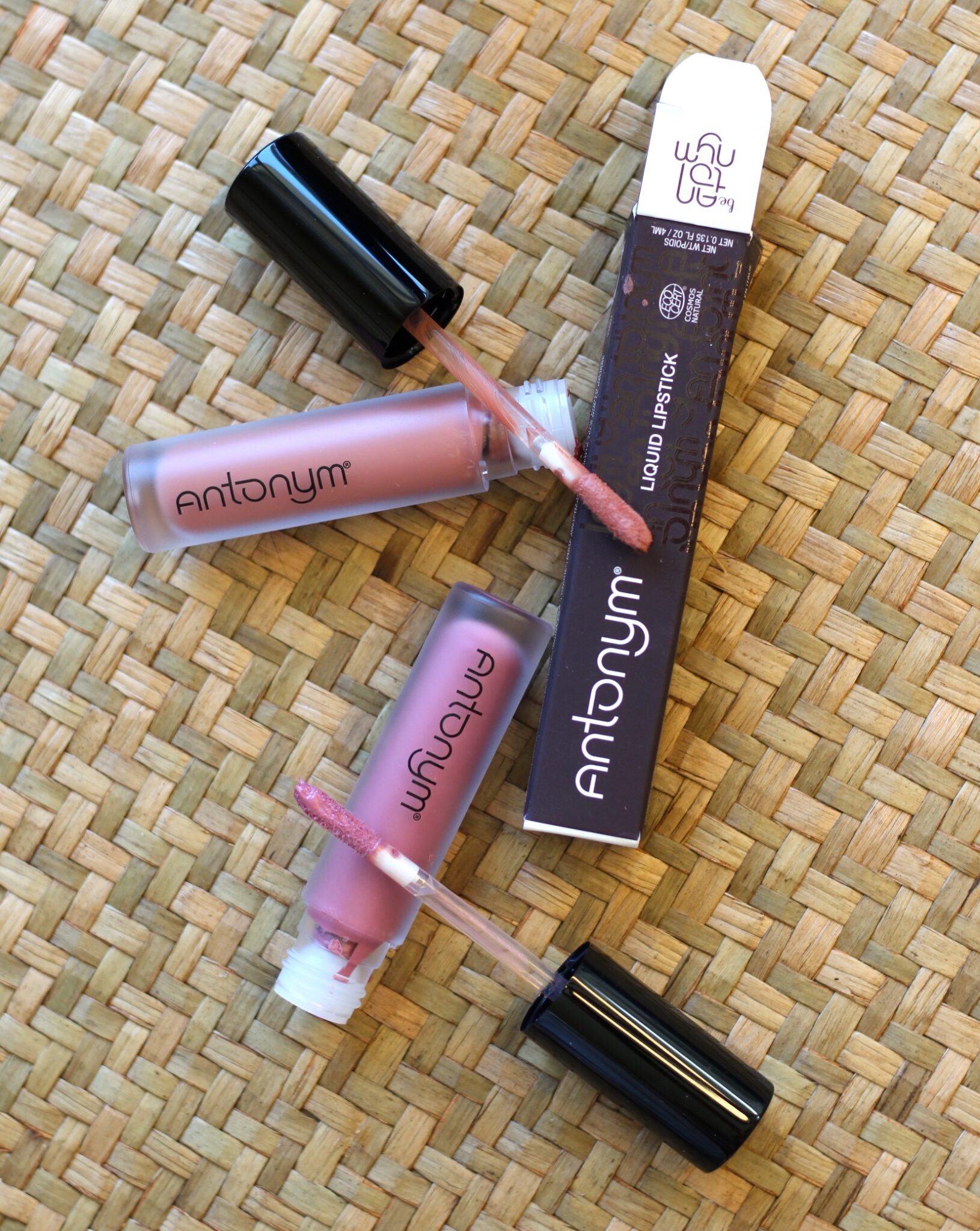 Two open tubes of Antonym liquid lipstick with applicators and a branded box are arranged on a woven surface.