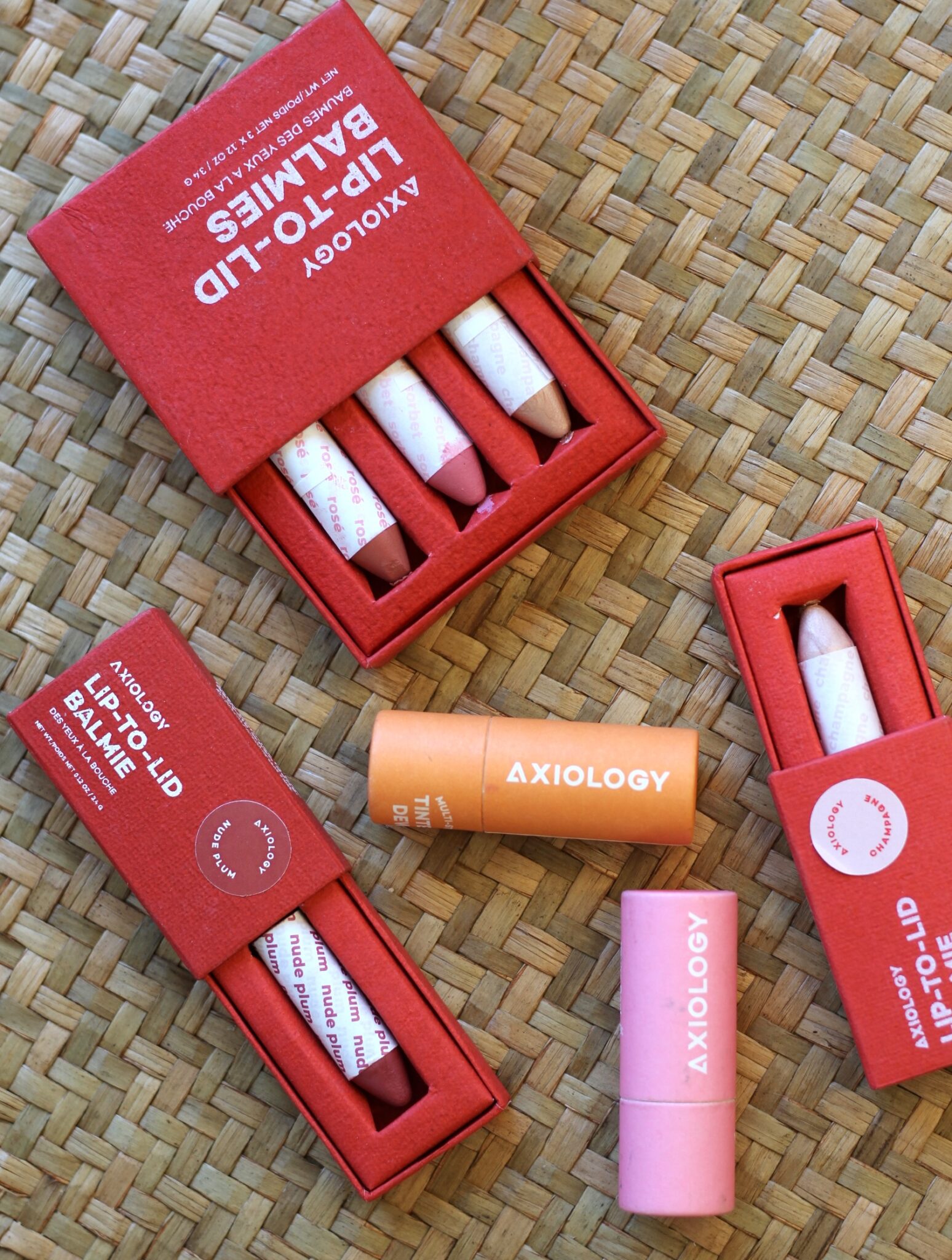 Several Axiology lip-to-lid balms in colorful, eco-friendly packaging are arranged on a woven surface.