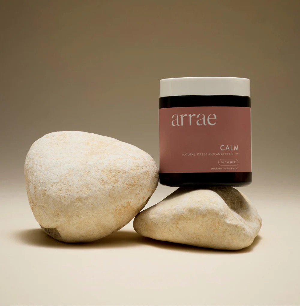 A jar labeled "arrae CALM" sits balanced on two smooth, beige rocks against a neutral background.