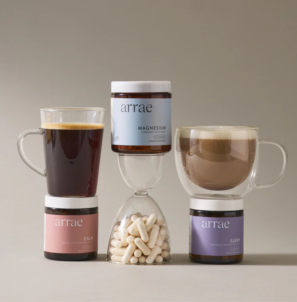 Two mugs of coffee flank an hourglass filled with capsules, with three Arrae supplement jars—Magnesium, Calm, and Sleep—arranged around the hourglass.