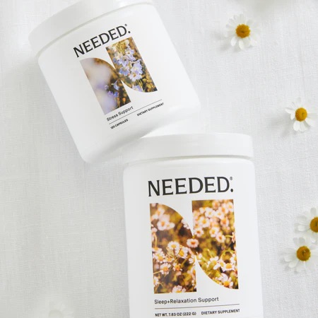 Two white containers labeled "NEEDED" on a white surface with small flowers; one is for stress support, the other for sleep-relaxation support.