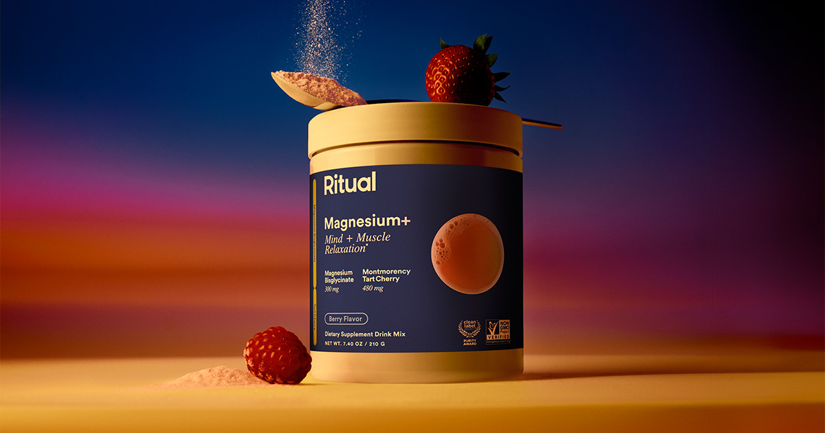 A container of Ritual Magnesium+ powder with a scoop of powder above it, strawberries on and around the lid, and powder scattered on the surface against a colorful background.