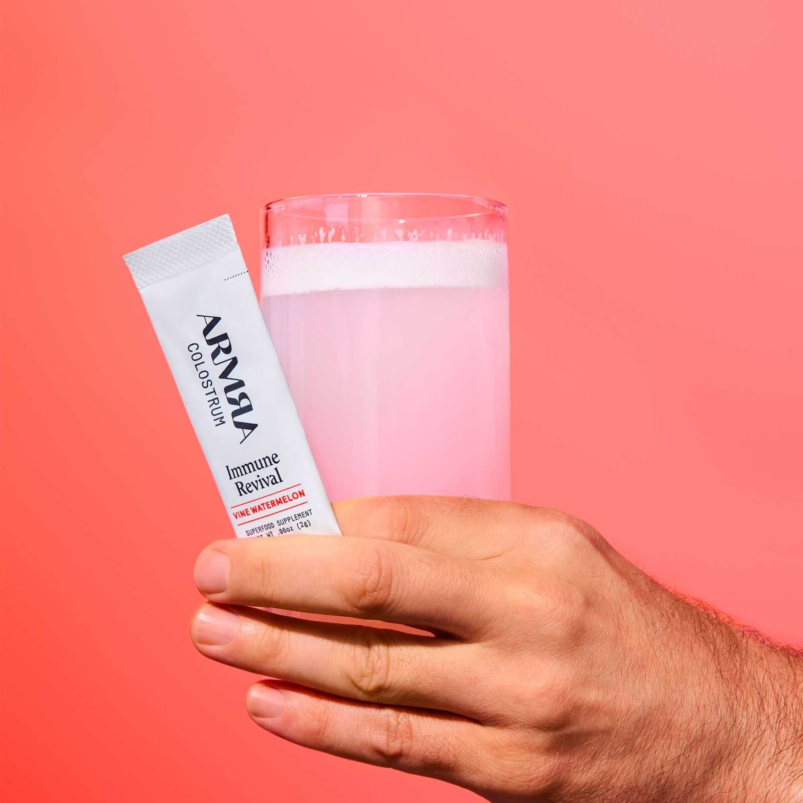 A hand holds a sachet labeled "ARMA Immune Revival" and a glass filled with a frothy pink beverage against a solid pink background.