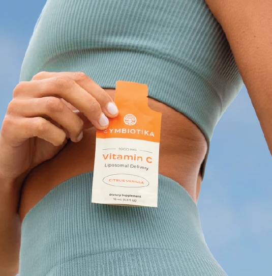A person in a blue ribbed athletic outfit holds a packet of Cymbiotika Vitamin C Liposomal Delivery supplement labeled Citrus Vanilla.