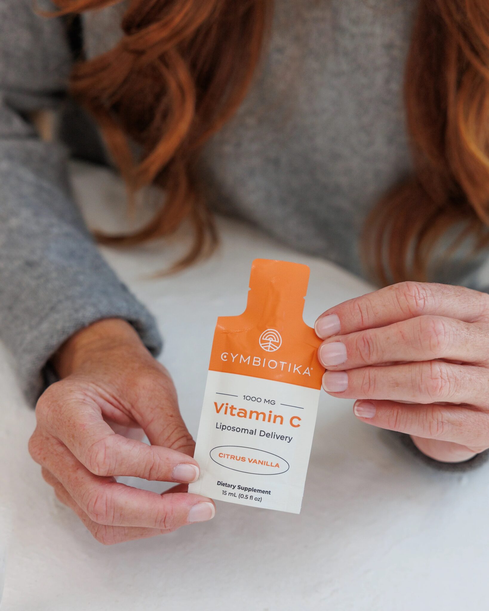 A person holds a packet of Cymbiotika Vitamin C Liposomal Delivery, Citrus Vanilla flavor, labeled as a dietary supplement with 1000 mg per serving.