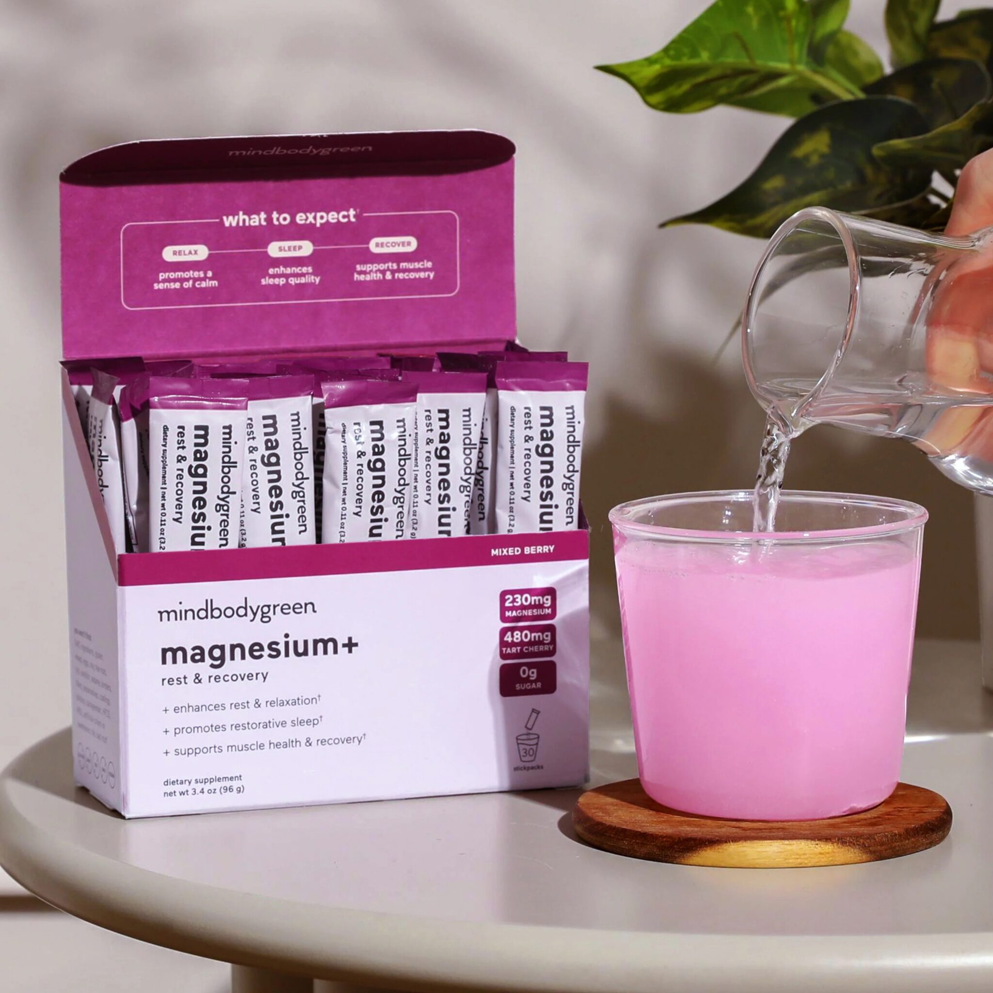 A hand pours water into a glass containing pink powder beside a box of mindbodygreen magnesium+ supplement packets on a table.
