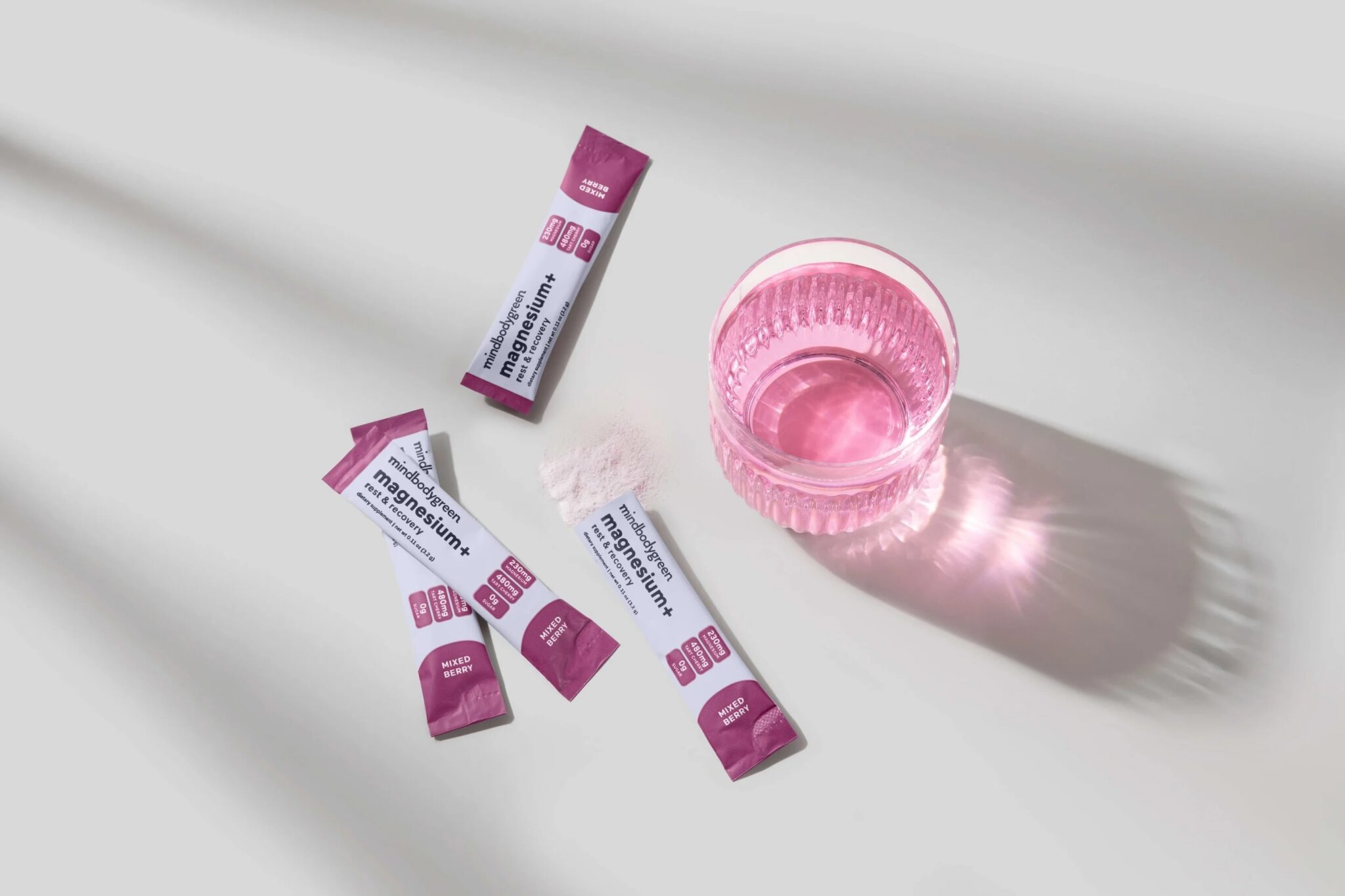 Three sachets labeled “magnesium” and one empty sachet are on a white surface next to a glass of pink liquid, with a small pile of white powder nearby.