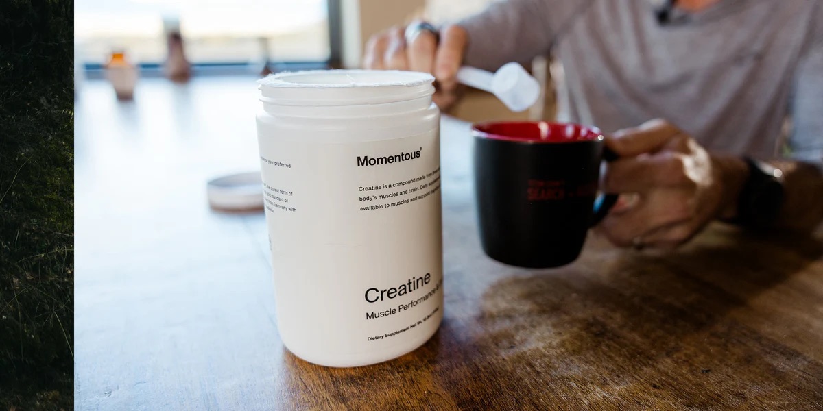 A person scoops powder from a container labeled "Momentous Creatine Muscle Performance" into a red and black mug on a wooden table.