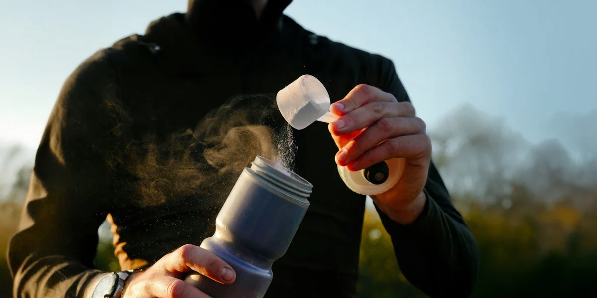 A person pours powdered supplement from a scoop into a sports bottle outdoors, with sunlight illuminating the powder.