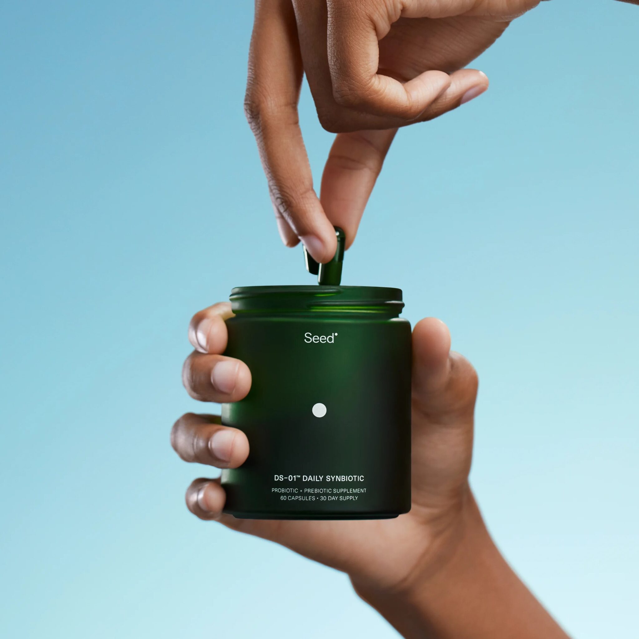 A hand holds a green Seed probiotic jar while another hand pinches and removes a capsule from the jar against a blue background.