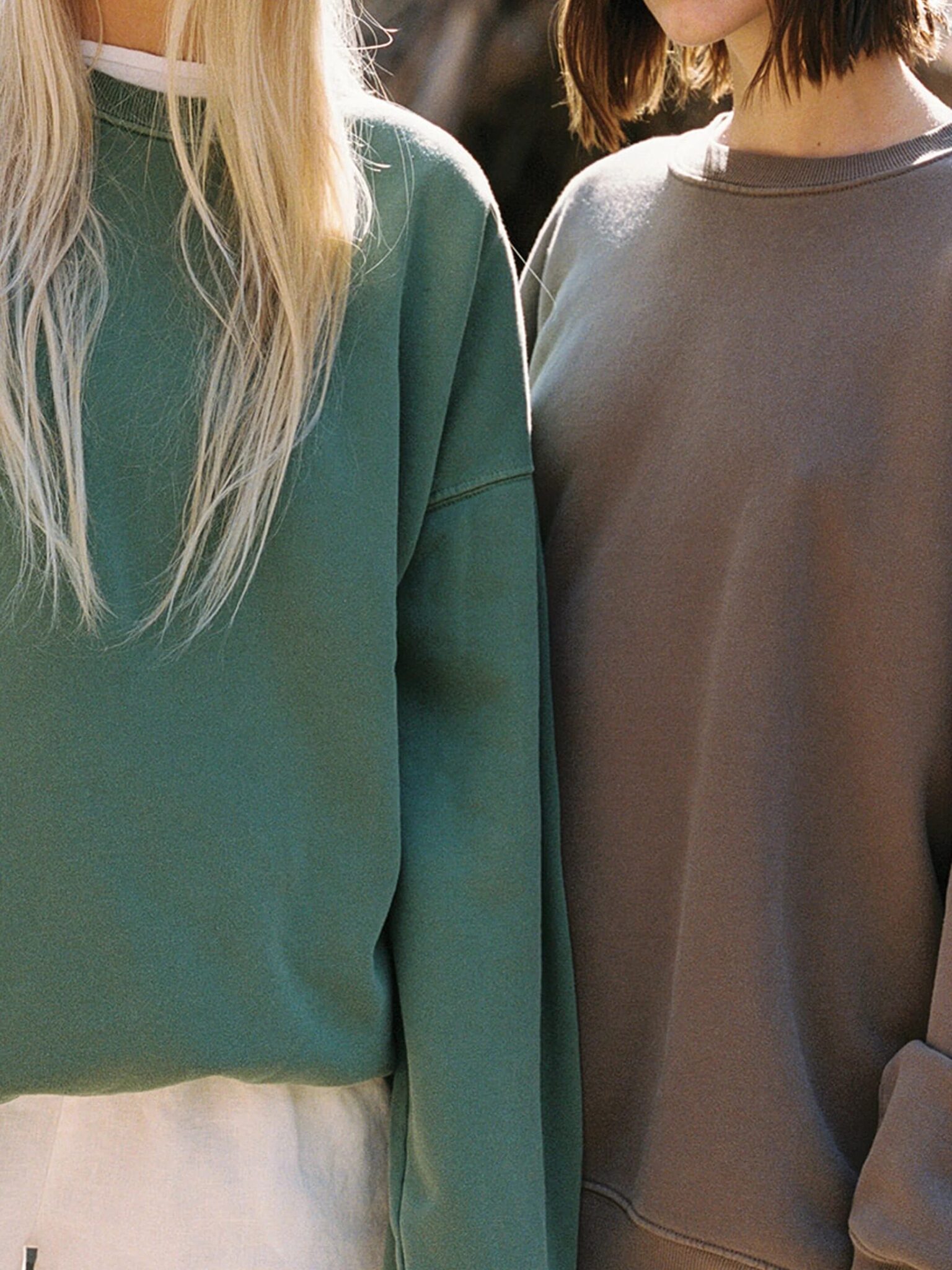 Two people standing side by side wearing casual crewneck sweatshirts, one green and one brown, with light-colored pants. Their faces are not fully visible.
