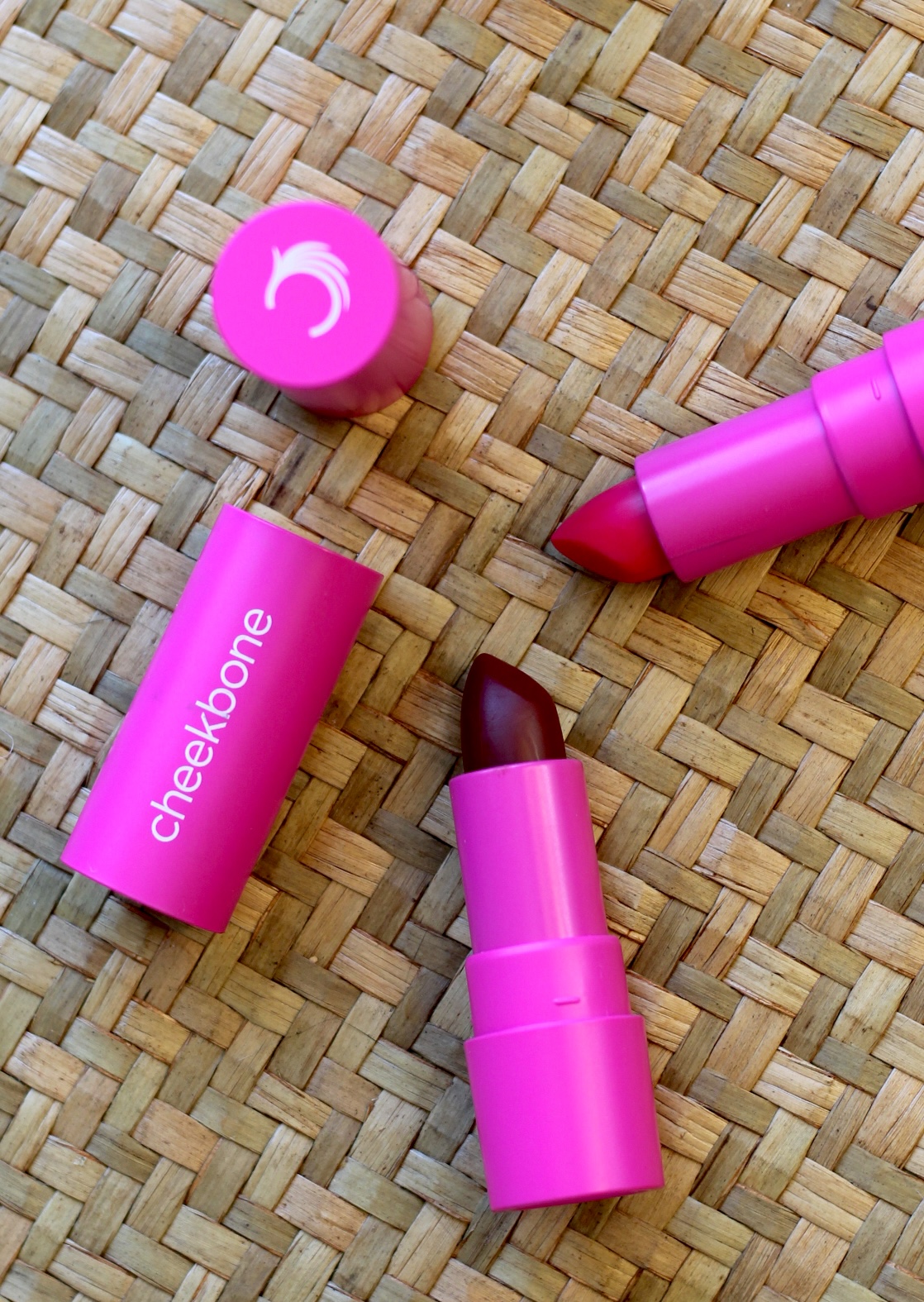 Two pink lipstick tubes, one open showing red lipstick and one partially closed, lie on a woven straw surface. The word "cheekbone" is printed on one tube.