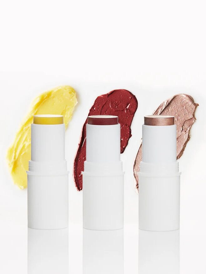 Three white tubes of makeup sticks in yellow, red, and bronze shades are displayed upright, each with a swatch of color behind them.