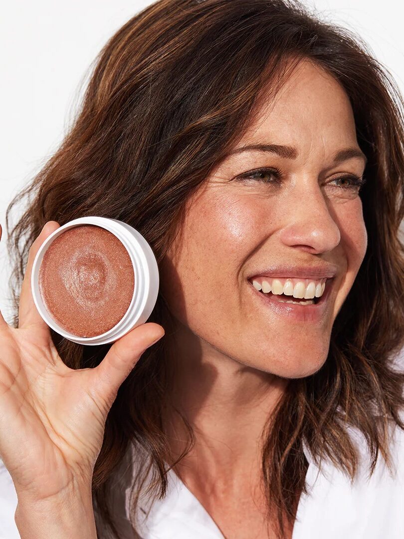 A woman with brown hair smiles while holding up an open container of shimmery bronzer against a white background.