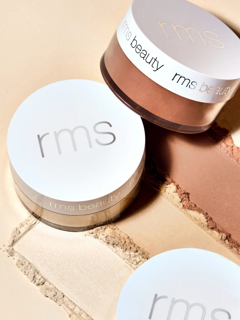 Three RMS Beauty makeup containers sit on a beige surface with makeup powders in light and bronze shades smeared beside each container.