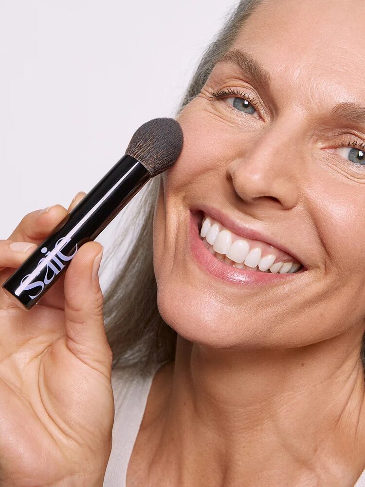 A woman smiles while applying makeup to her cheek with a brush.