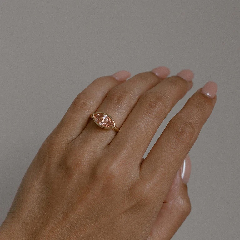 A hand with light pink manicured nails wears a gold ring featuring a marquise-shaped design with two small stones on the ring finger.