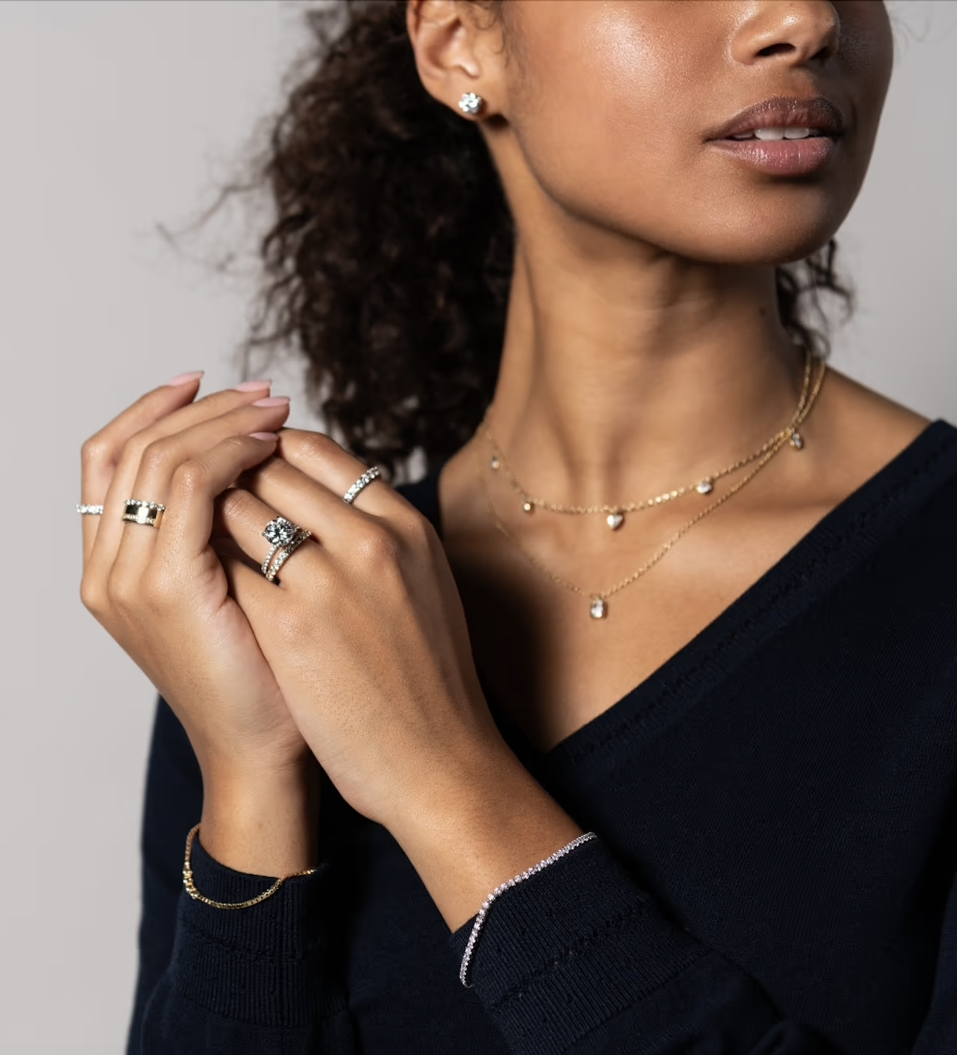 A woman wearing layered gold necklaces, multiple rings, a bracelet, and stud earrings, dressed in a dark V-neck top.