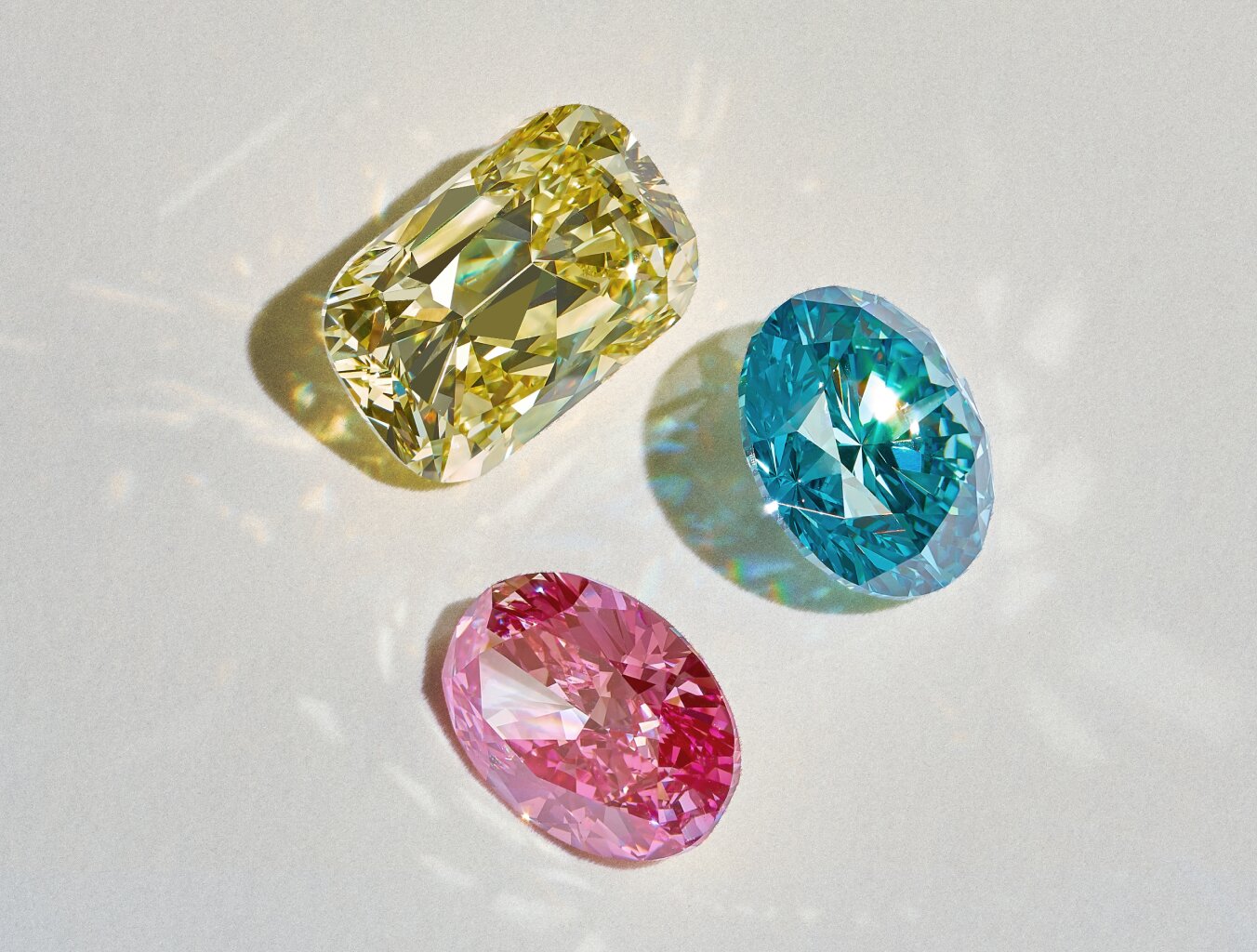 Three faceted gemstones—one yellow, one blue, and one pink—are displayed on a light surface, with visible reflections and colorful light patterns.