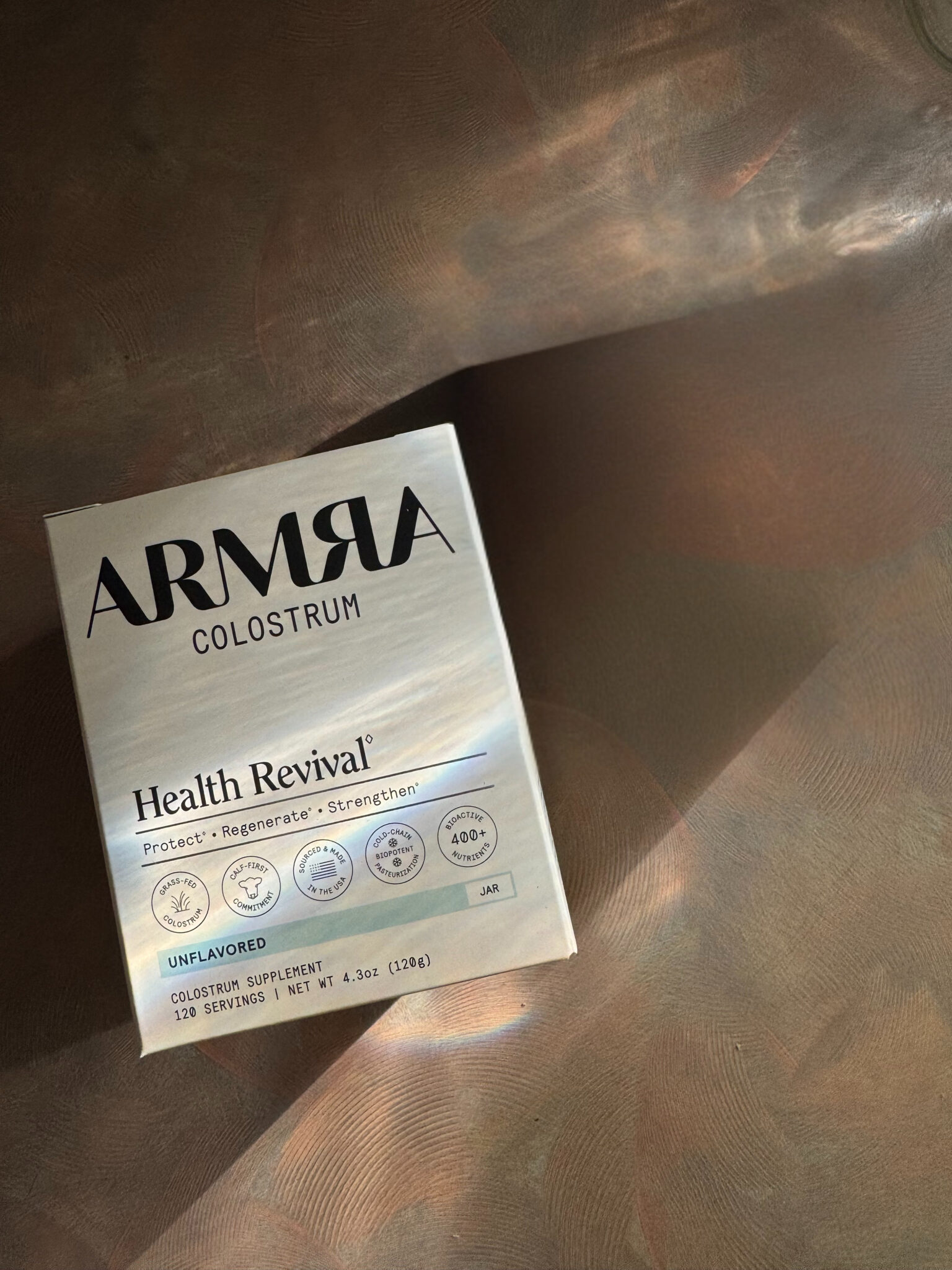 Box of Armra Colostrum Health Revival supplement in unflavored variety, photographed on a metallic, textured surface with light reflections.