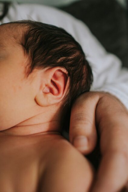 Close-up of a person's hand gently resting on a baby's upper back, showing the baby's ear and side of the head in soft natural light.