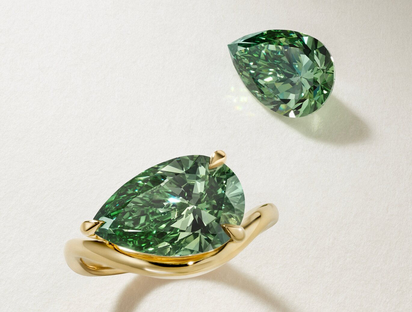 A gold ring with a pear-shaped green gemstone is displayed next to a loose, similarly cut green gemstone on a light surface.