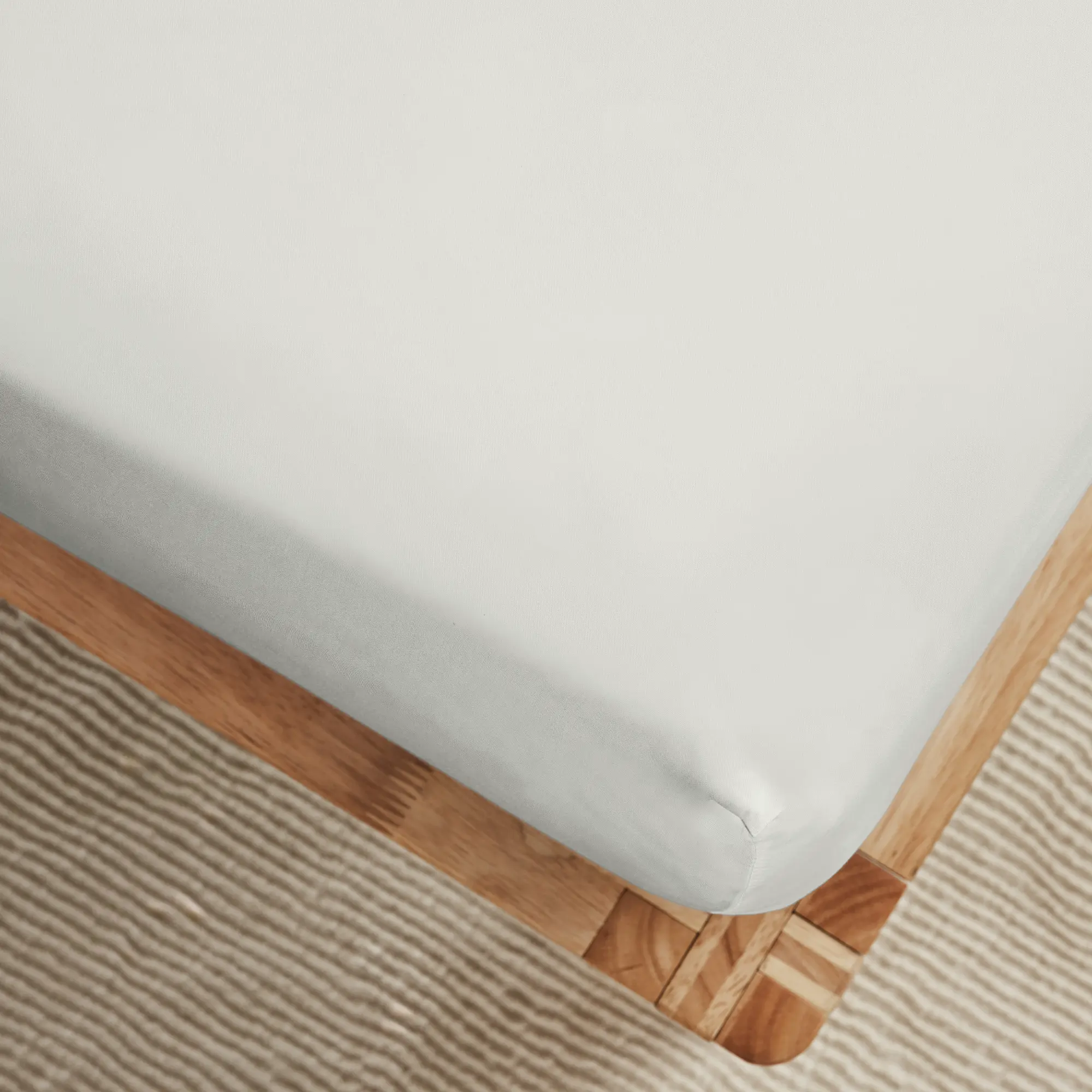 Close-up of a corner of a bed with a white fitted sheet on a wooden frame, with a textured beige rug underneath.