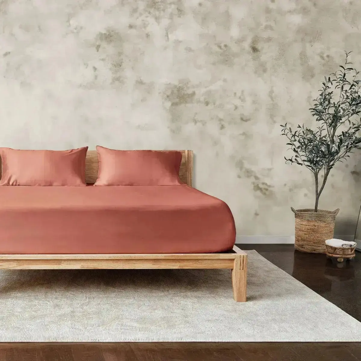 A minimalist bedroom features a wooden bed with rust-colored bedding, a textured beige wall, a woven basket with a potted plant, and a light rug on a dark wood floor.