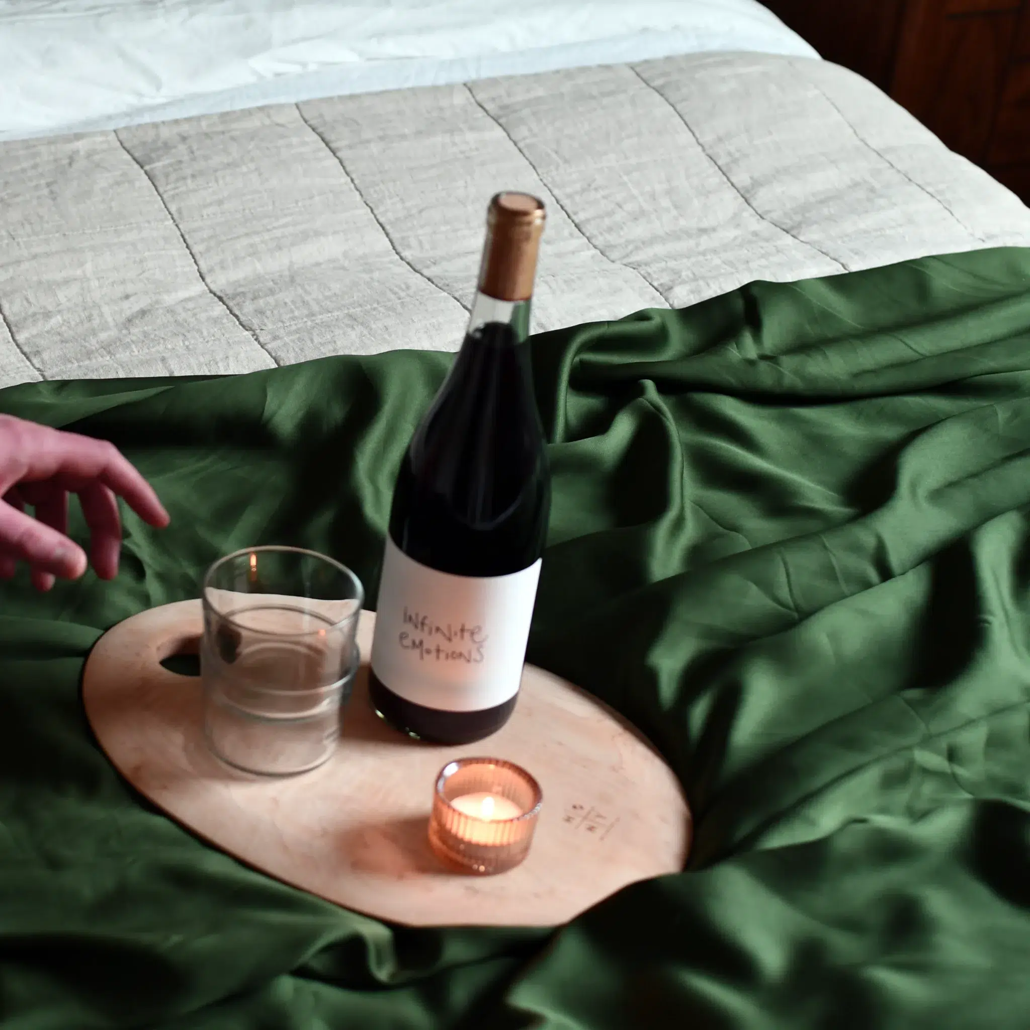 A wooden tray on a green bedspread holds a wine bottle labeled "Infinite Emotions," a glass of water, and a lit tealight candle; a hand reaches toward the tray.