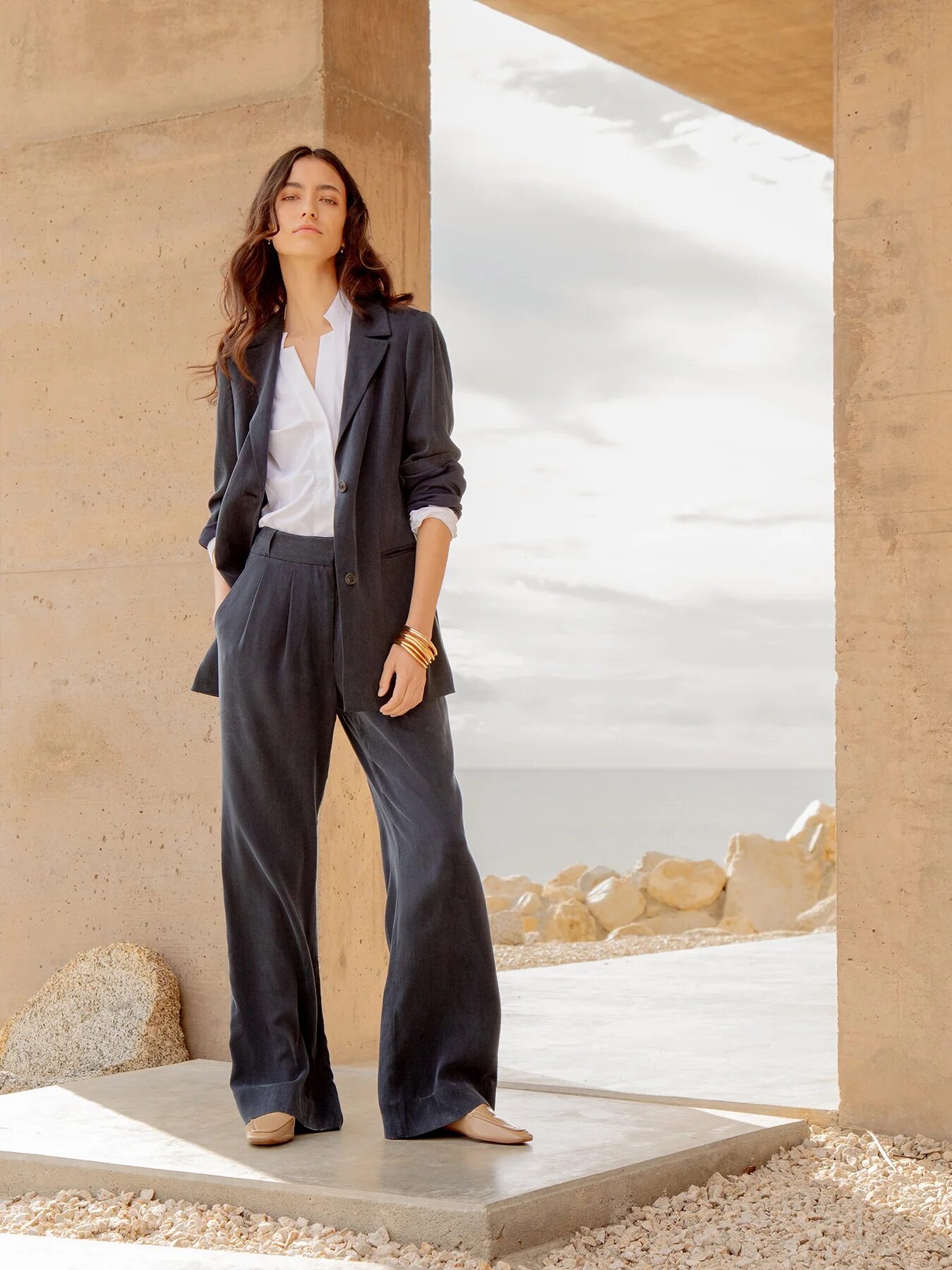 A woman stands outdoors near a concrete structure, wearing a loose-fitting dark suit and white shirt, with rocks and ocean in the background.