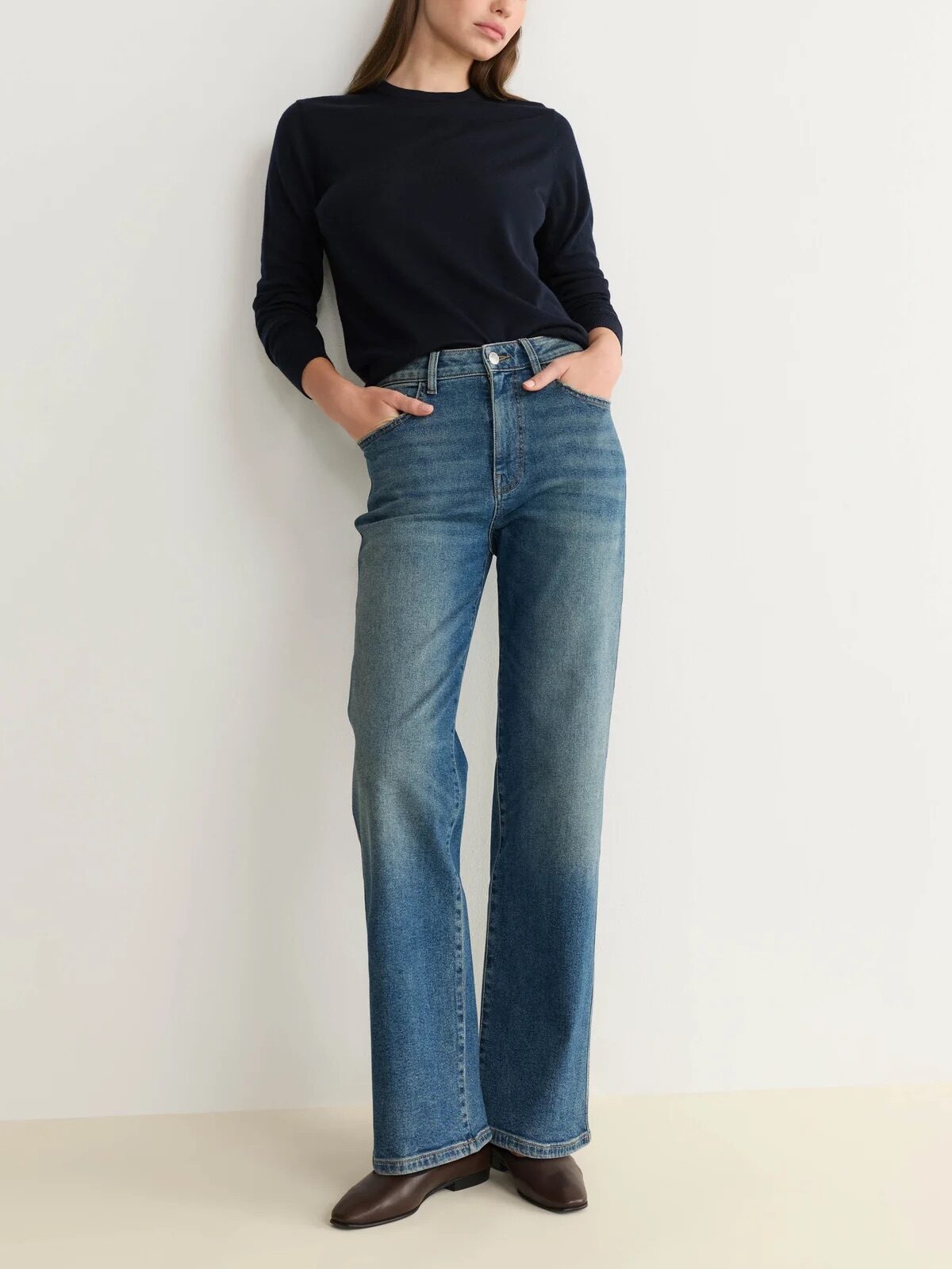 Woman standing against a plain wall wearing a black long-sleeve top, blue high-waisted jeans, and brown shoes with her hands in her pockets.