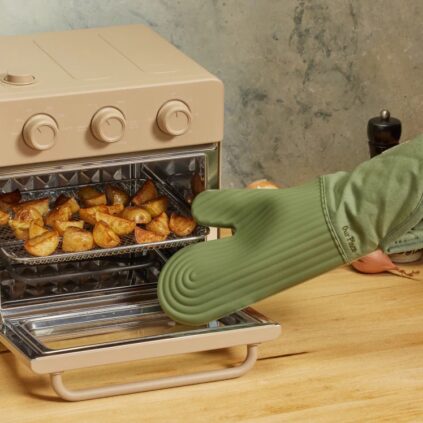 A hand wearing a green oven mitt pulls a tray of roasted potato wedges out of a beige countertop oven.