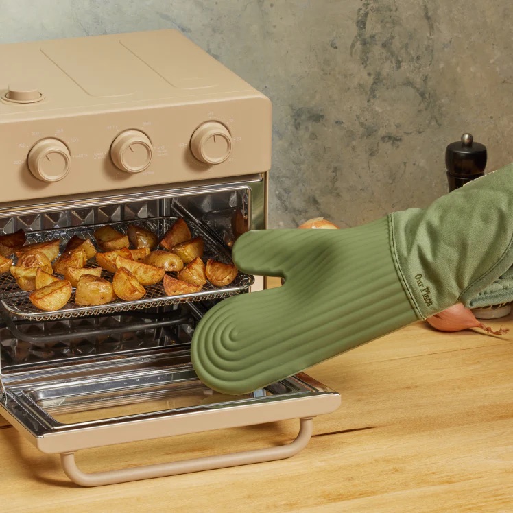A hand wearing a green oven mitt pulls a tray of roasted potato wedges out of a beige countertop oven.