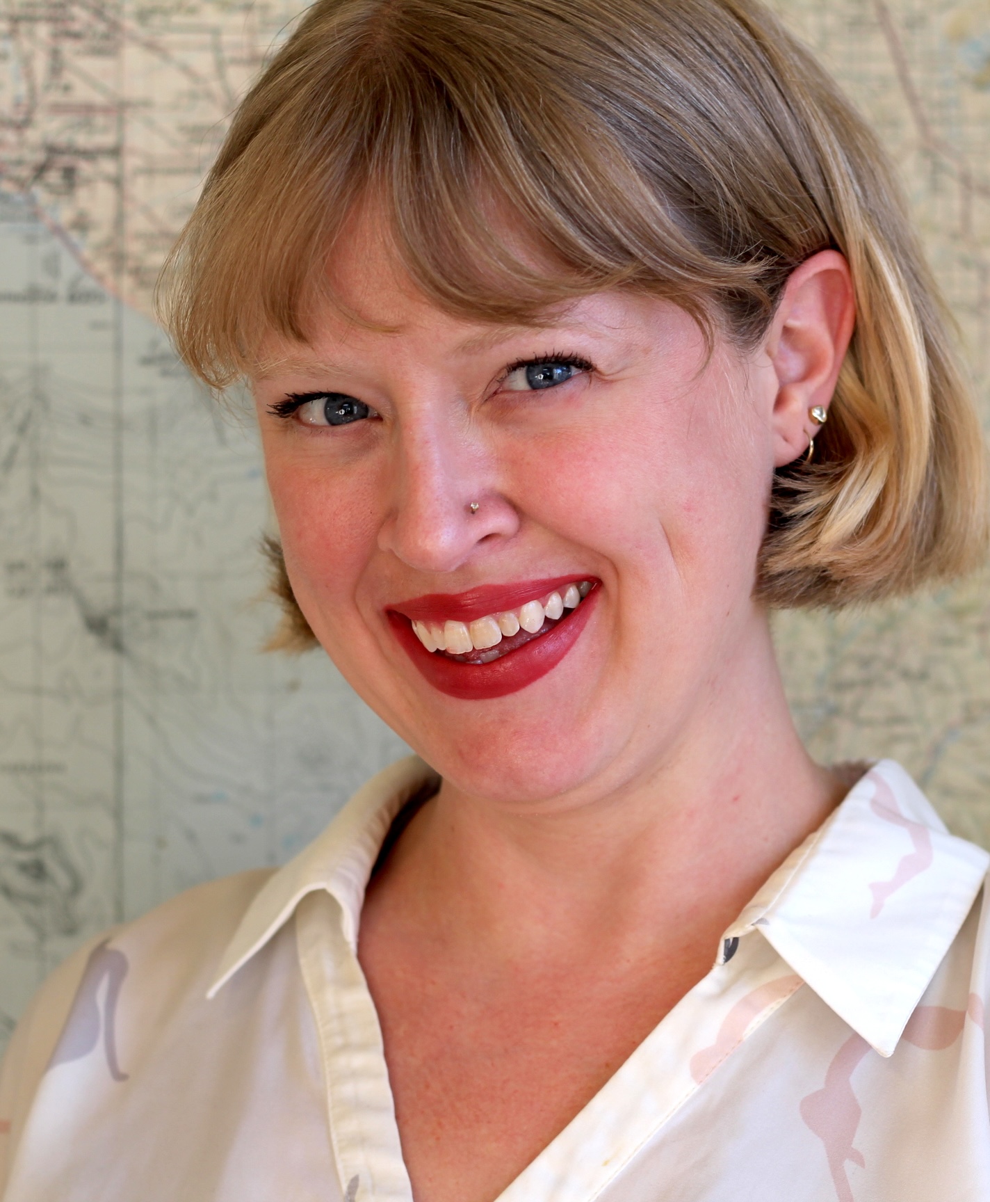 A woman with short blonde hair, wearing a white blouse, smiles at the camera. A map is visible in the background.
