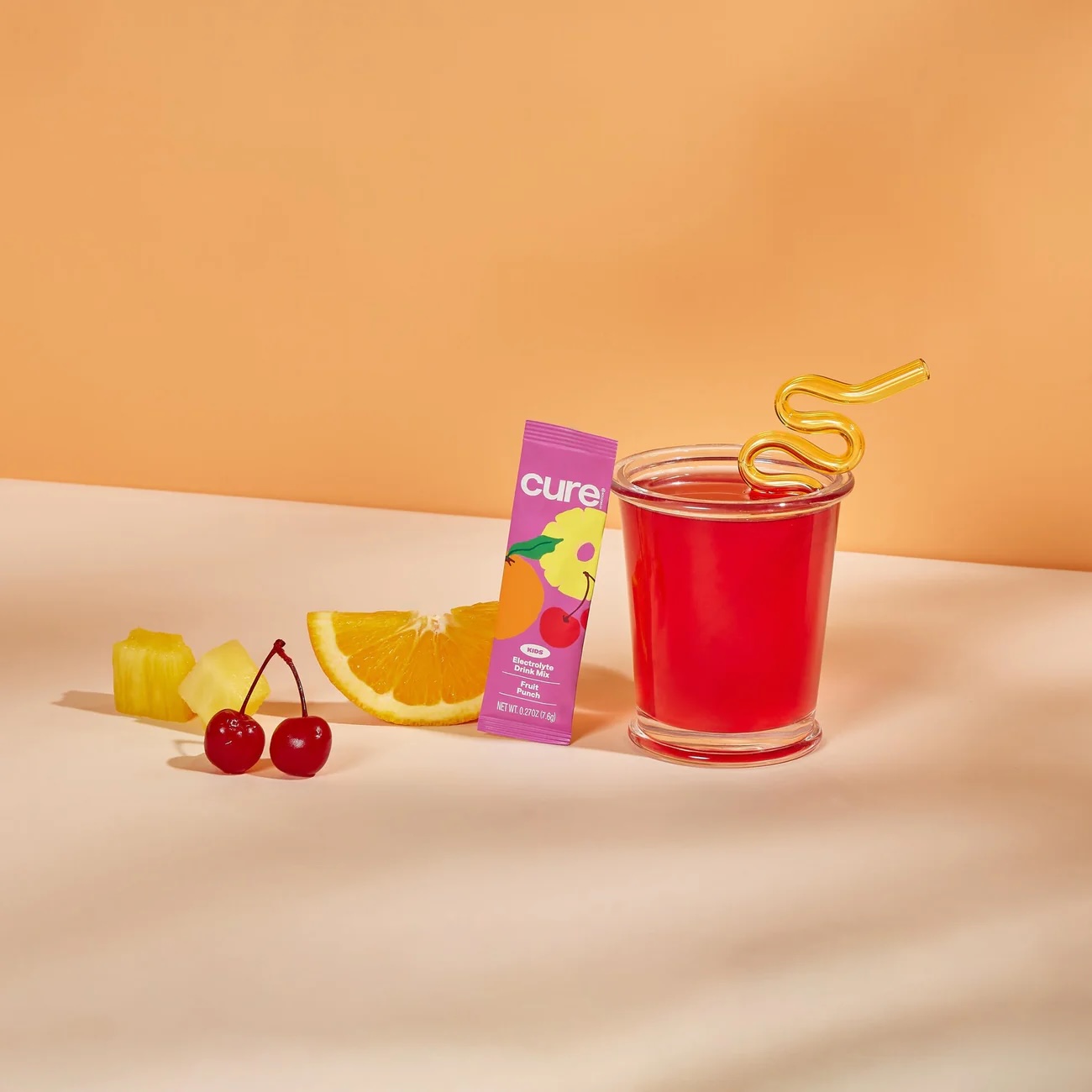 A glass of red drink with a curly straw sits next to a Cure Hydration packet, pineapple pieces, orange slice, and two cherries on a beige surface with an orange background.