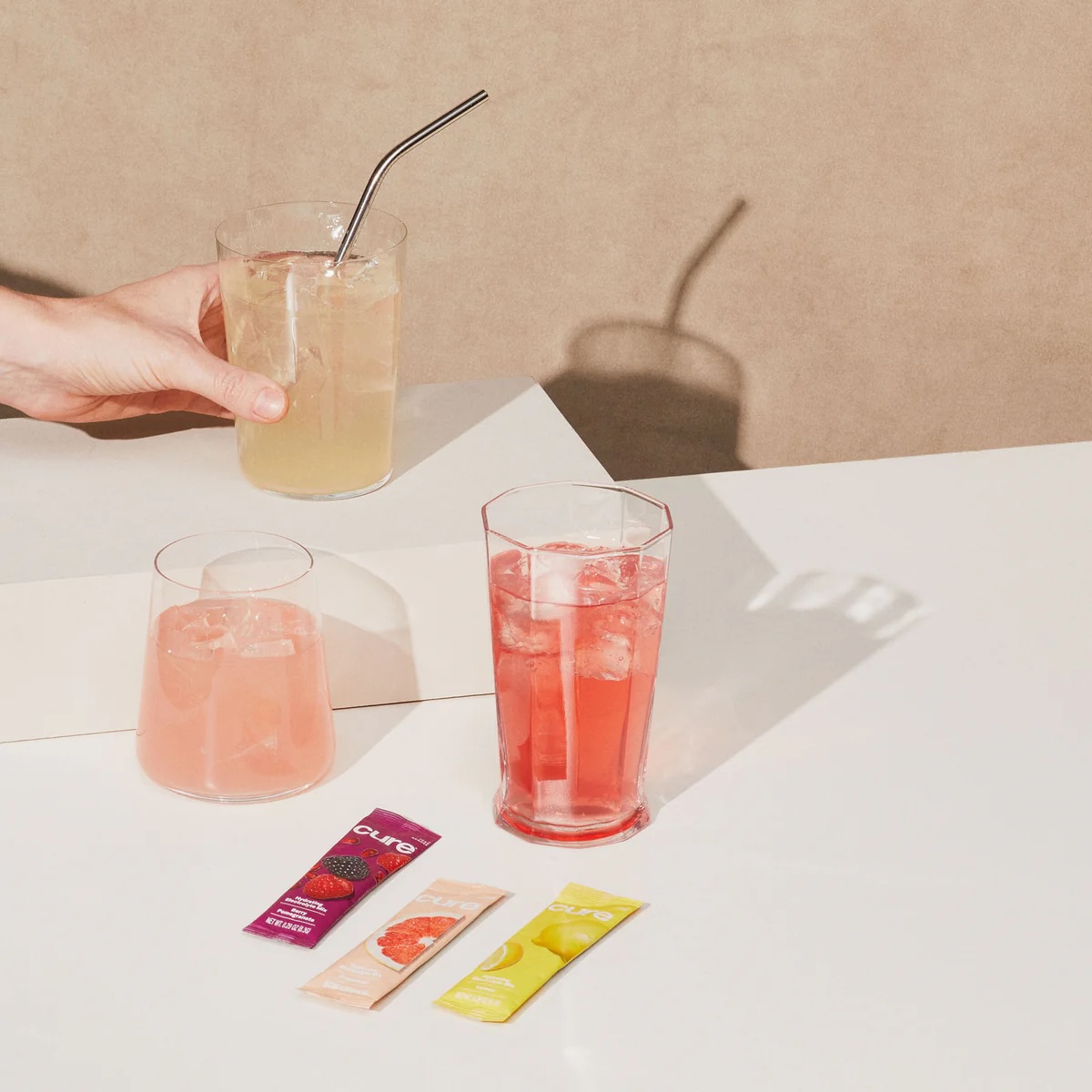 Two glasses of pink and red drinks with ice are on a white table next to three drink mix packets; a hand holds a glass with a metal straw in the background.